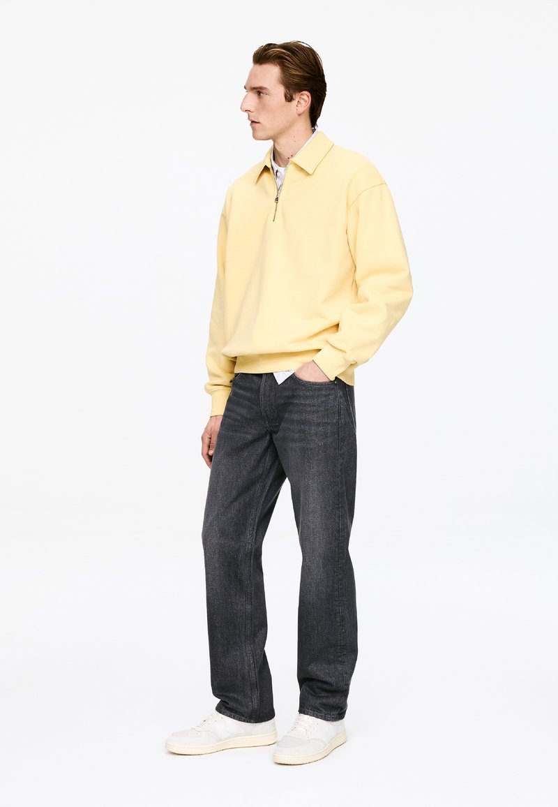 Pull jaune clair à fermeture éclair, jean gris et baskets blanches. Le pull présente un col et une coupe décontractée, tandis que le denim a une finition légèrement texturée.