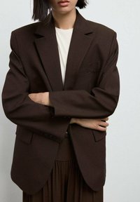 Blazer oversize marrone con spalle strutturate, una tasca e due bottoni. Indossato sopra un top crema, il tessuto appare liscio e strutturato.