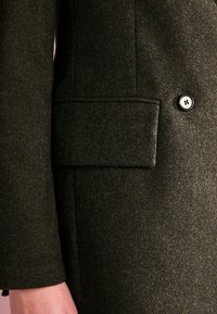 Mørkegrøn uldjakke med en sidelomme og en sort knap. Stoffet har en blød tekstur med et subtilt, fuzzy udseende.