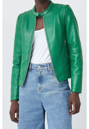 Personne portant une veste en cuir verte ajustée par-dessus une chemise blanche et un jean bleu clair taille haute, debout devant un fond uni.