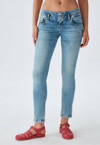 Jean skinny en denim bleu clair avec une taille haute, cinq poches et une fermeture à deux boutons. Porté avec des chaussures rouges à lanières.