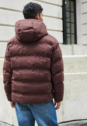 Jachetă pufosică maro cu glugă, design pătratul cu matlasare, și manșete elastice. Asortată cu blugi din denim bleu deschis.