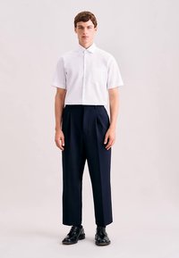 Weißes kurzärmeliges Hemd mit Tasche, kombiniert mit marineblauen, hoch taillierten plissierten Hosen und schwarzen Schuhen. Schlichter Hintergrund.