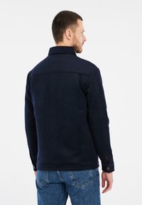 Tamsiai mėlyna striukė pagaminta iš storos medžiagos, su apykakle, sagų rankovėmis ir tiesiu kirpimu. Matomos siūlės gale.