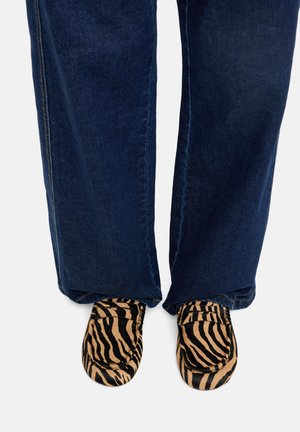 Osoba ubrana w ciemnoniebieskie dżinsy i mokasyny w nadruk zebry, stojąca na jasnym tle.