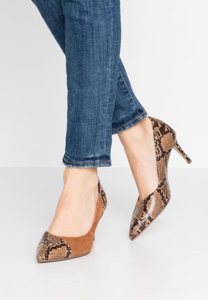 Fødder i blå jeans iført brune og slangeskindsmønstrede spidse højhælede sko, den ene fod krydset en smule foran den anden.