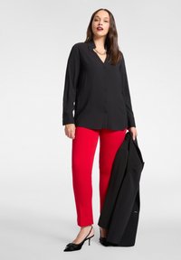 Czarna koszula na guziki, czerwone obcisłe spodnie, czarne sandały na obcasie z kokardką, trzyma czarną marynarkę. Gładka tkanina, dopasowany krój, żywe kolory.