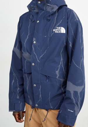 Marineblaue North Face wasserdichte Jacke mit Blitzmuster, Knöpfen vorne, verstellbarer Kapuze und sichtbarer beigefarbener Hose darunter.