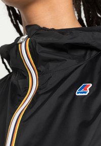 Schwarze Windjacke mit hohem Kragen, ausgestattet mit einem mehrfarbigen Reißverschluss, silbernen Beschlägen und einem Logo-Patch auf der Brust.