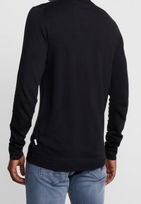 Schwarzes Langarmoberteil mit taillierter Passform, gerippten Bündchen und einem schlichten Halsausschnitt, kombiniert mit hellblauen Jeans. Etikett am Saum.