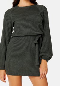Femme portant une robe pull côtelée vert olive avec de longues manches bouffantes et une ceinture nouée sur le côté, posant les bras détendus.