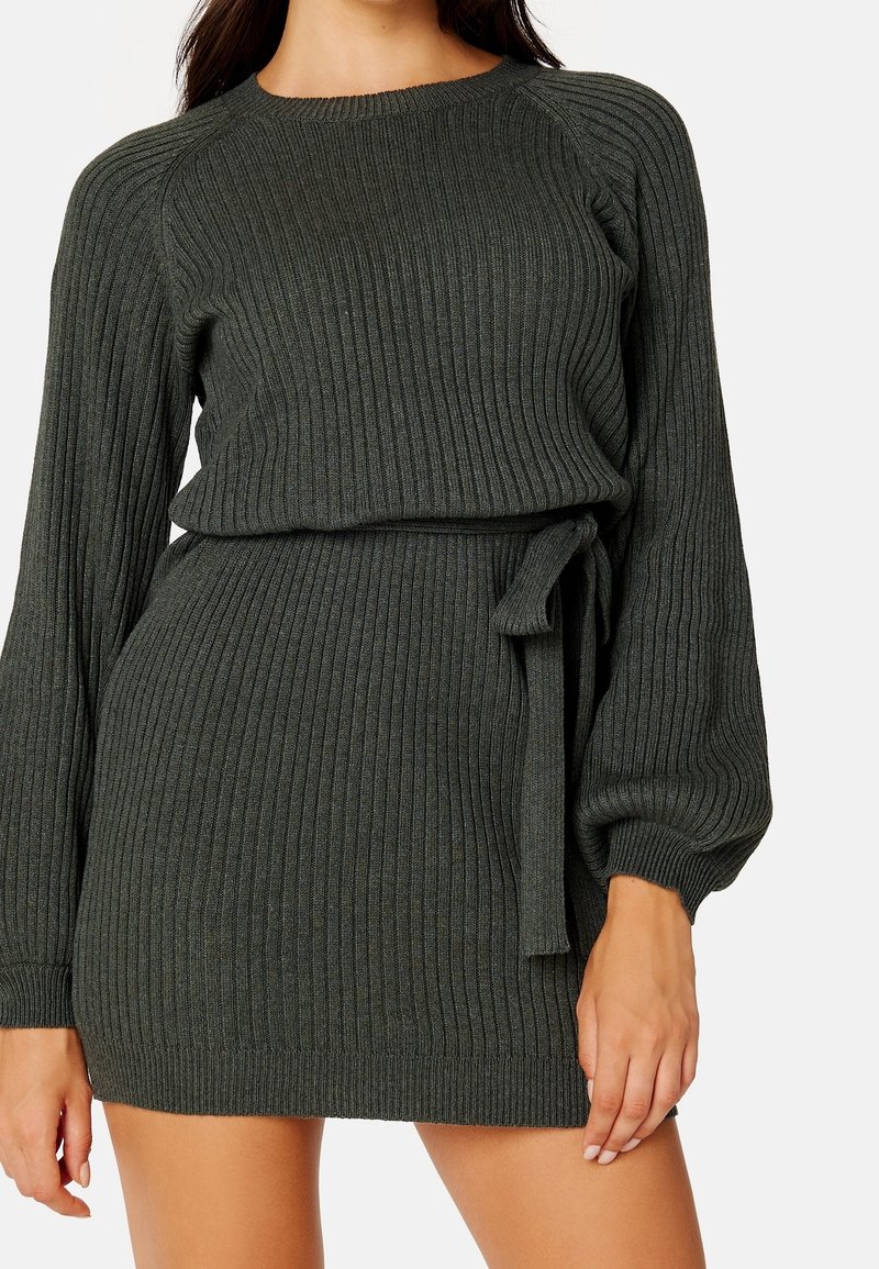Femme portant une robe pull côtelée vert olive avec de longues manches bouffantes et une ceinture nouée sur le côté, posant les bras détendus.