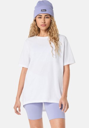 Weißes, übergroßes T-Shirt mit kurzen Ärmeln, kombiniert mit helllila Fahrradshorts. Das Modell trägt eine helllila Mütze mit schwarzem Logetikett.