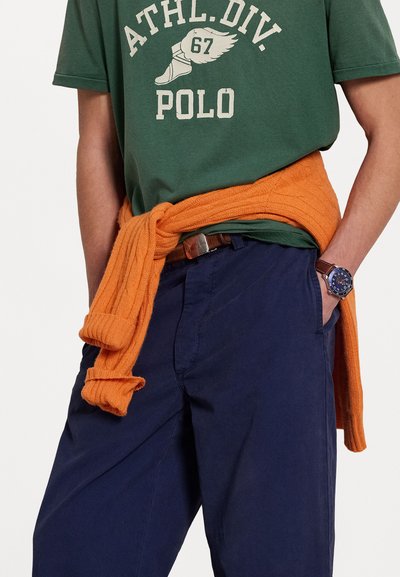 T-shirt en coton vert avec texte imprimé blanc, pull tricoté orange noué autour de la taille, et pantalon bleu marine. Ceinture brune et montre.