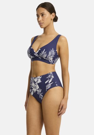 Das Model trägt ein marineblaues Blumenbikini-Set, das aus einem Wickel-Oberteil und einer hochgeschnittenen Unterhose besteht. Sie posiert selbstbewusst mit einem neutralen Gesichtsausdruck.