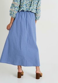 Falda maxi a rayas azul y blanca con cintura elástica, tejido fluido y corte holgado, combinada con sandalias de tacón cuadrado marrones.