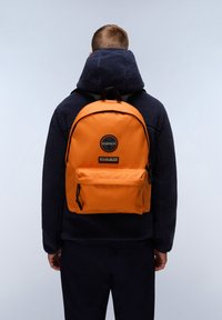 Zaino arancione in nylon con forma arrotondata, tasca anteriore e una patch rotonda nera sullo sopra. Indossato sopra una giacca in fleece scura.