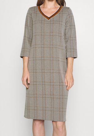 Kobieta ubrana w sukienkę w kratę sięgającą kolan, z rękawami do 3/4 długości oraz dekoltem w kształcie litery V wykończonym na brązowo i czarno.