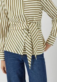 Gestreepte shirt in crèmekleur en olijfgroen, met een wikkelontwerp, strik in de taille, knoopdetails en lange mouwen. Gecombineerd met blauwe jeans.