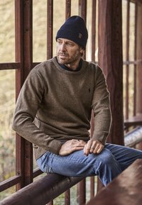 Bruine wollen trui met geribbelde manchetten en hals, spijkerbroek en een blauwe gebreide muts, zittend bij houten balken en ramen.