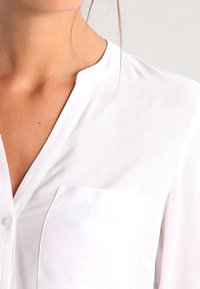 Blusa bianca in tessuto morbido, con colletto a risvolto, tasca frontale e dettagli in bottone sulla pattina. Texture liscia e aspetto leggero.