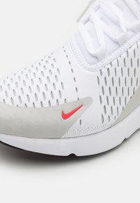 Vita idrottssko med en strukturerad ovandel, perforerad design, ljusgrå detaljer och en röd Nike swoosh-logotyp på sidan.