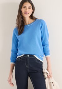 Pull léger bleu avec un col et des poignets côtelés. Associé à un jean foncé avec des coutures contrastantes, porté avec un manteau de couleur claire.