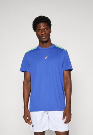 Man in een blauwe sport T-shirt met groene schouderaccenten en witte sportshorts die tegen een effen lichte achtergrond staat.