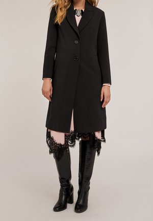 Femme portant un manteau noir boutonné jusqu'aux genoux sur une robe rose avec une bordure en dentelle noire et des bottes noires à talons hauts.