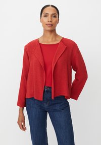 Rote strukturierte Jacke mit offenem Frontdesign, ohne Kragen und mit drei Viertel langen Ärmeln, getragen über einem roten Oberteil und blauen Jeans.