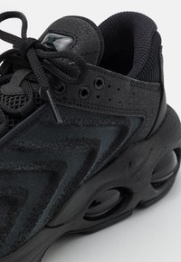 Gros plan sur une chaussure de sport noire avec tissu texturé, détails en mesh, lacets ronds et semelle épaisse avec des coussinets d'air visibles.