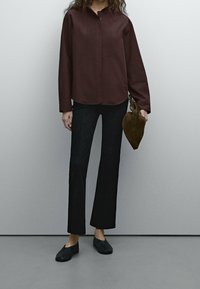 Chemise boutonnée bordeaux avec des rayures texturées, associée à un pantalon noir évasé et des chaussures noires. Tenant un sac pochette vert foncé.