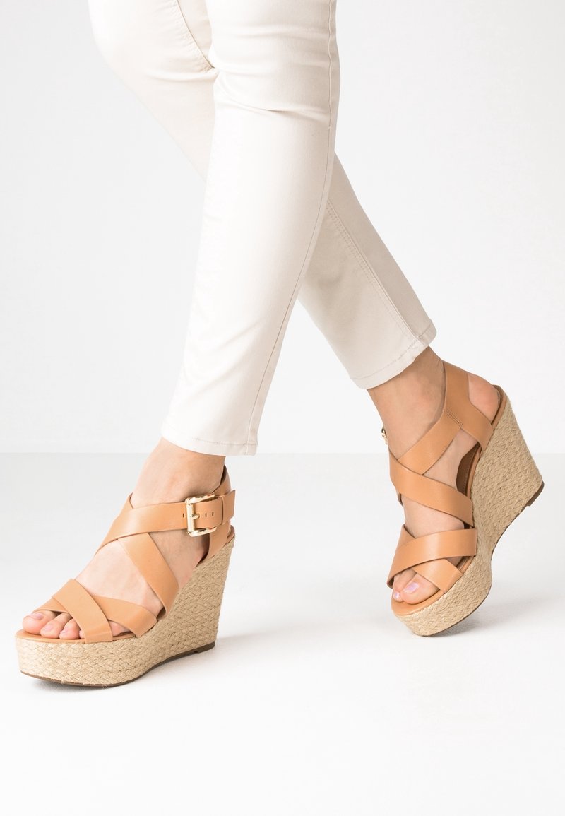 Sandalias de cuña de cuero marrón con correas cruzadas y hebilla en el tobillo, que cuentan con una suela de plataforma tejida texturizada y un diseño de punta abierta.