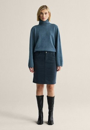 Kvinde iført en blå rullekrave, mørkt, knælængde denimskørt, sorte knæhøje støvler, stående mod en ensfarvet baggrund.