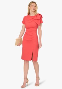 Koralowa sukienka o dopasowanym kroju, z krótkimi rękawami, dużą kokardą na jednym ramieniu i rozcięciem sięgającym uda. Złota kopertówka i sandałki na cienkich paskach.