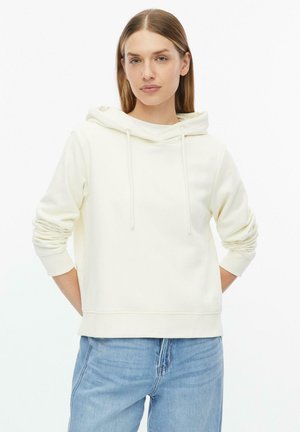 Frau mit glattem hellbraunem Haar, die einen weißen Kapuzenpullover und blaue Jeans trägt und mit den Händen hinter dem Rücken steht.