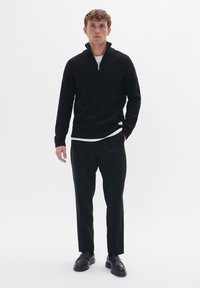 Suéter negro acanalado con cuello de media cremallera, llevado sobre una camisa blanca, combinado con pantalones negros y zapatos negros. Diseño simple, tejido texturizado.
