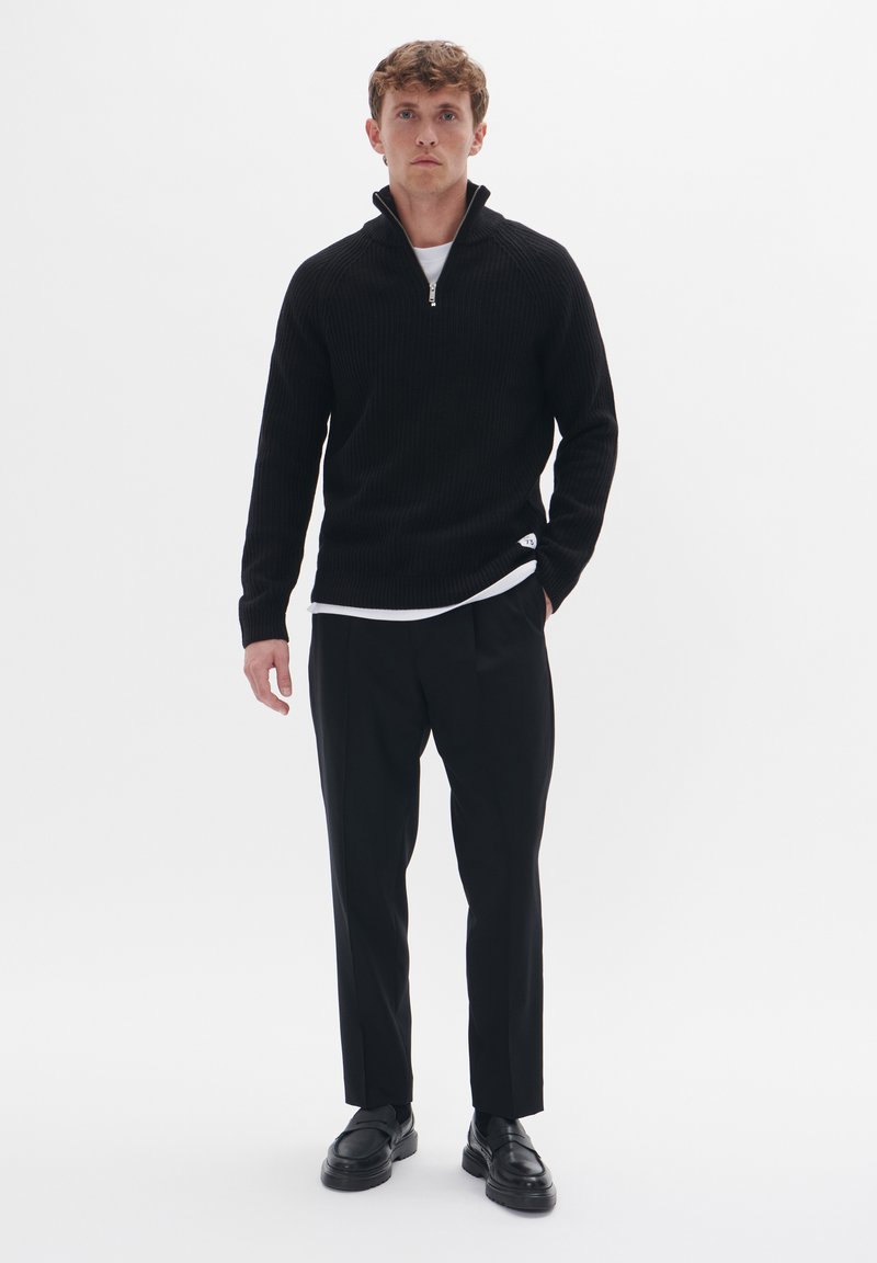 Suéter negro acanalado con cuello de media cremallera, llevado sobre una camisa blanca, combinado con pantalones negros y zapatos negros. Diseño simple, tejido texturizado.
