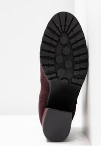 Semelle d’une botte de cheville bordeaux présentant une semelle noire épaisse avec des motifs hexagonaux et linéaires, et un talon carré texturé.
