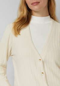 Rippenstrick-Cardigan in Cremefarbe mit tiefem V-Ausschnitt und drei Knöpfen. Mit langen Ärmeln und weichem, strukturiertem Stoff.