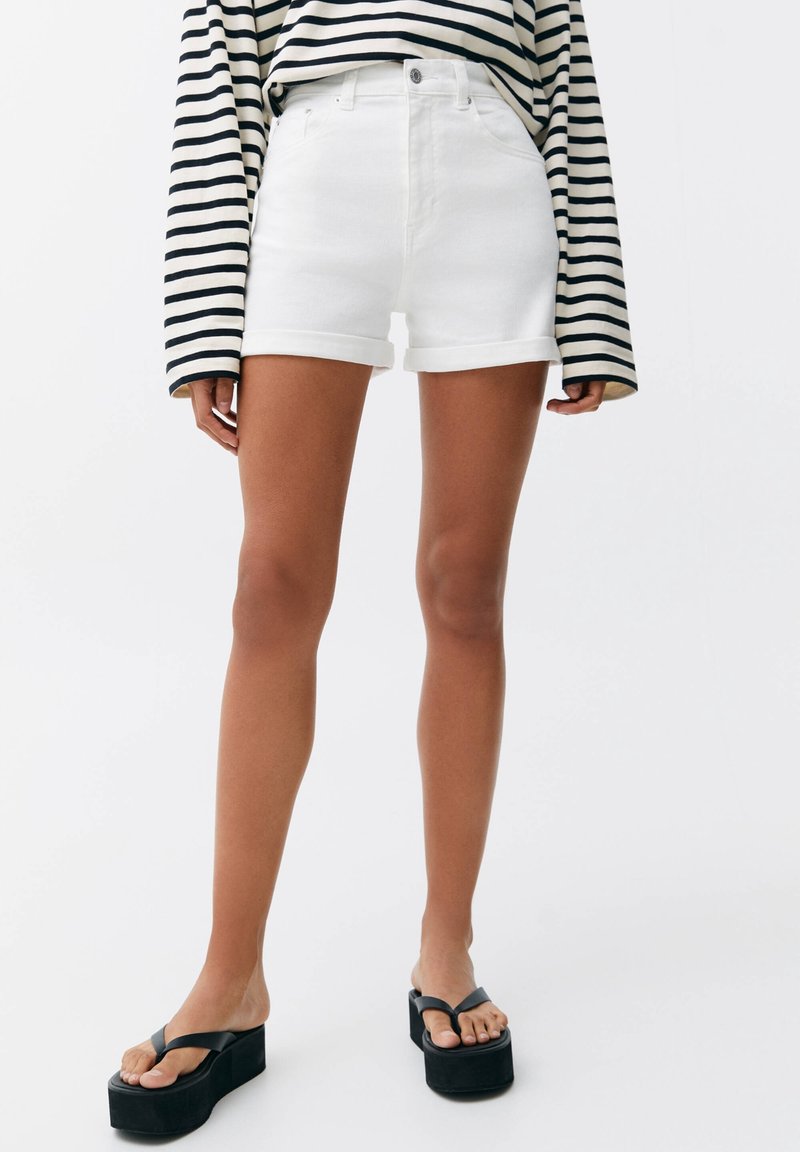 Femme portant un short en denim blanc à ourlet roulé, des tongs à plateforme noires et un chemisier à manches longues rayé noir et blanc.