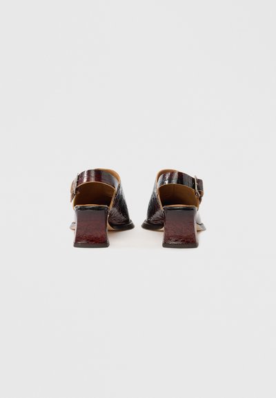 Zadaj pogled na mule z visokim petom iz sijočega bordo materiala. Ponašajo se z debelim petom, odprtim zadnjim delom in nastavljivim trakom z zaponko.