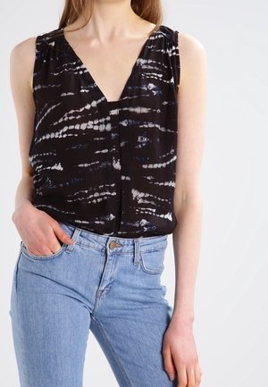 Kobieta w czarnej bezrękawnikowej bluzce tie-dye z dekoltem w serek, w połączeniu z jasnoniebieskimi, wysokim stanem jeansami, jedna ręka w kieszeni.