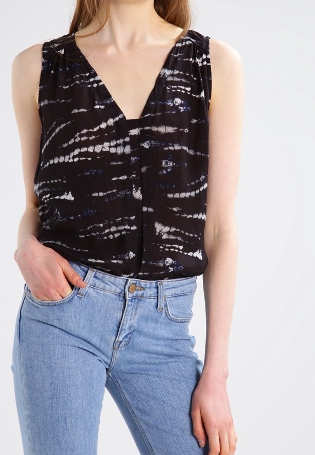 Femme portant un chemisier sans manches noir tie-dye avec un col en V, associé à un jean taille haute bleu clair, une main dans la poche.