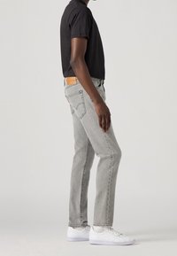 Jasnoszare dżinsy o prostym kroju nogawki, z delikatnym przetarciem i opaloną skórzaną naszywką z tyłu, w zestawieniu z czarnym t-shirtem z krótkim rękawem i białymi trampkami.