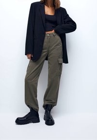 Veste noire cintrée portée sur un crop top foncé, associée à un pantalon cargo vert olive avec poches latérales, et des bottines noires épaisses.