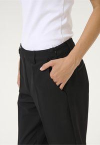 Pantalon noir à texture lisse, doté de poches avant, d'une fermeture éclair et d'une taille ajustée, associé à un haut blanc.