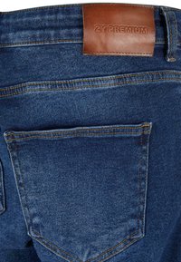 Temno modri jeans iz denima z gladko teksturo, ki imajo rjavo usnjeno oznako z napisom "2Y PREMIUM" in eno zadnjo žepono.