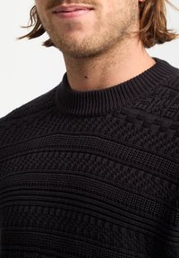 Sort strikket sweater med et tekstureret mønster, der har en ribbet rund hals og vandrette striber. Nærbillede af kraveområdet.