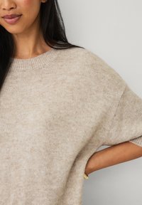 Beiger gestrickter Pullover mit weitem Halsausschnitt und fallenden Schultern. Weiche Textur, subtile Sprenkel und lockerer Schnitt. Sichtbare Hand in der Taschen-Detail.